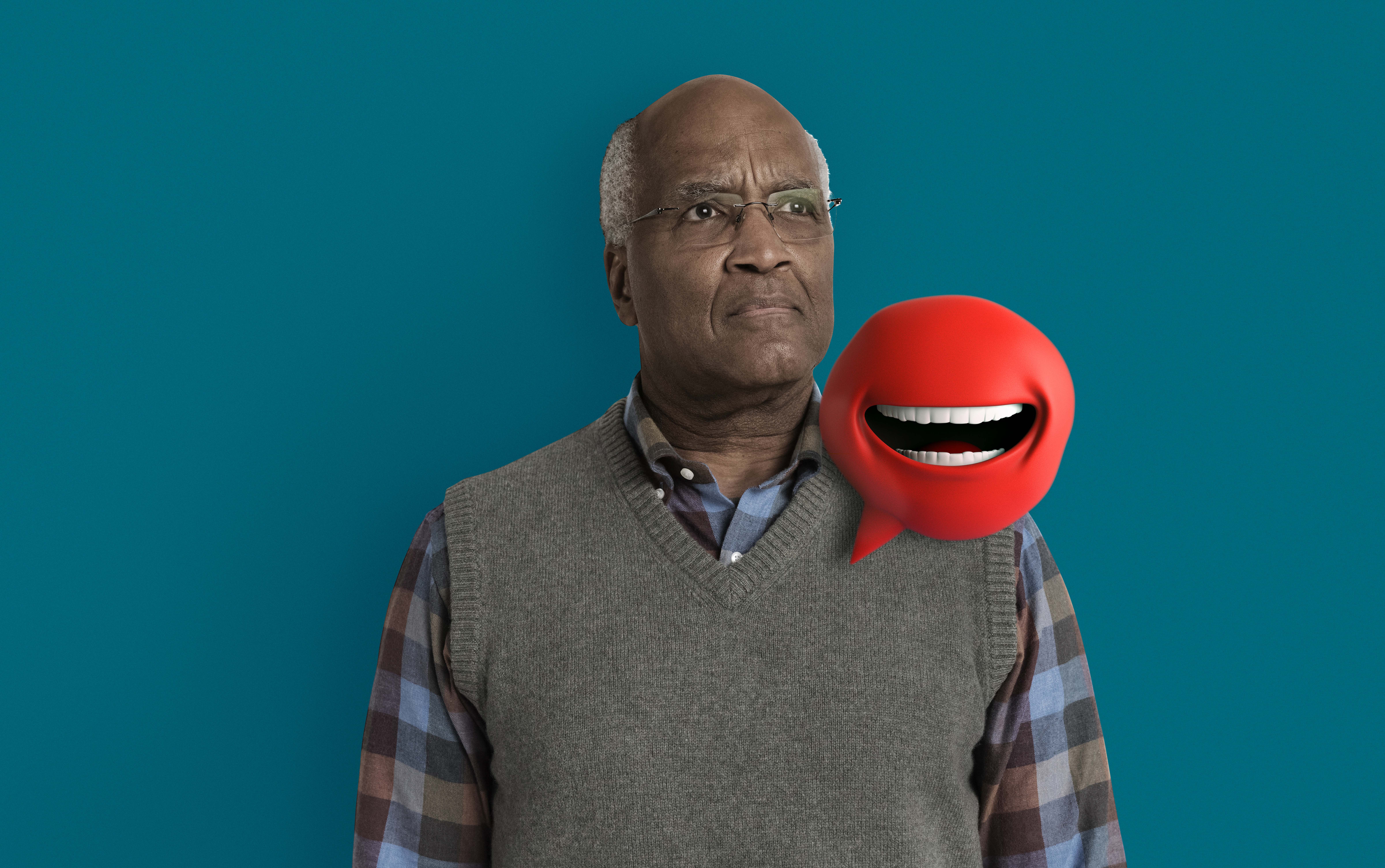 Heart screening health campaign visual A black man in his 60s, with glasses, a tweed pull over and plaid shirt stands looking out to the middle distance, with a red, rounded 3D speech mark character smiles to camera. It has no eyes, a mouth, and a grin.